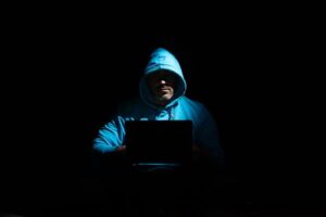 A person in a hoodie using a laptop in a dark setting, conveying mystery and technology themes.